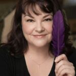 Woman with dark hair holding a purple feather close to her face, smiling, wearing a black top and a delicate necklace.