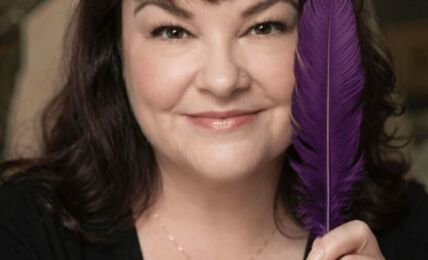 Woman with dark hair holding a purple feather close to her face, smiling, wearing a black top and a delicate necklace.