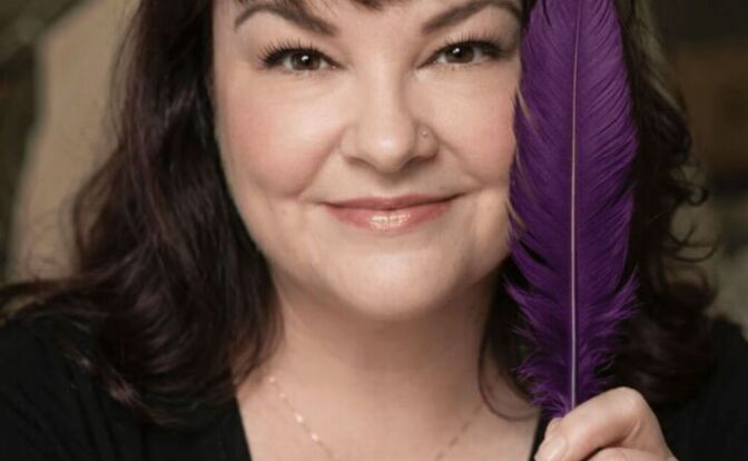 Woman with dark hair holding a purple feather close to her face, smiling, wearing a black top and a delicate necklace.